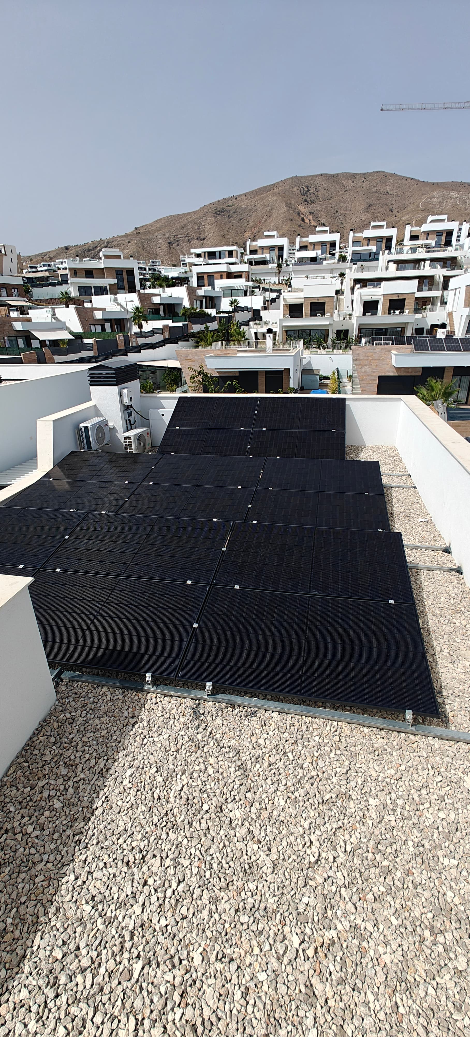 Paneles solares negros sobre azotea de grava con casas blancas y una montaña al fondo.
