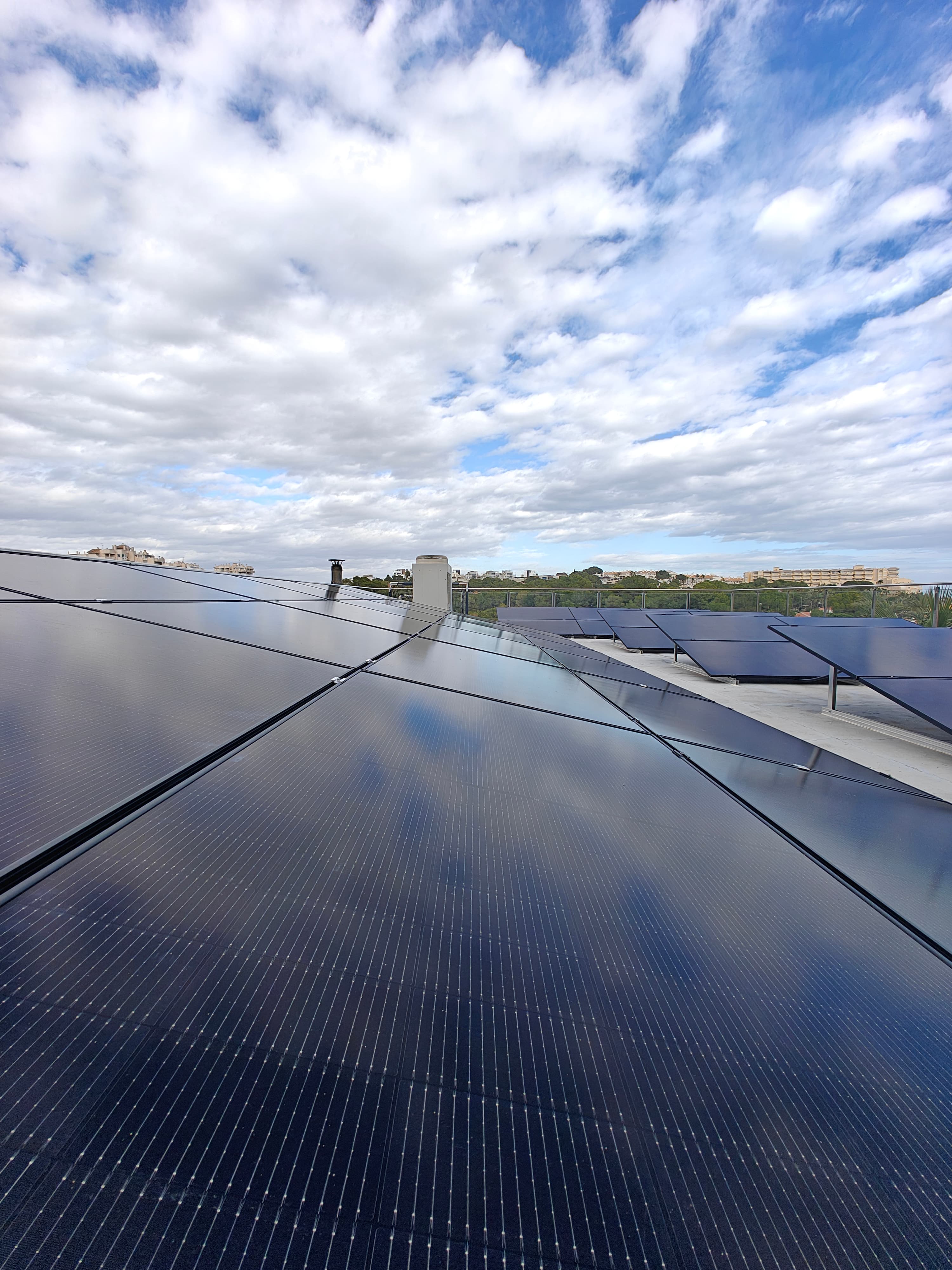 Paneles solares en una azotea reflejan un cielo nublado sobre un paisaje urbano distante.