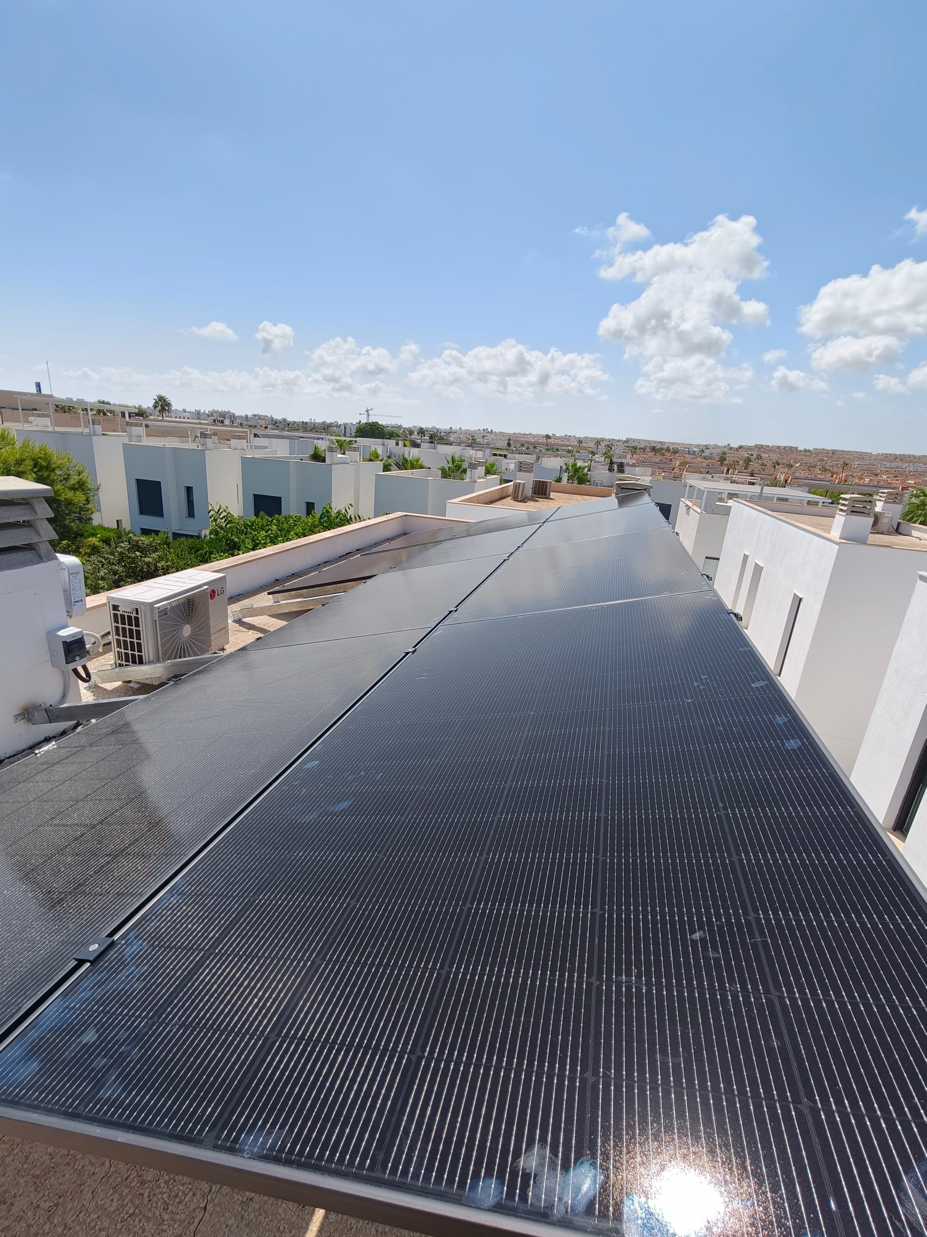 Paneles solares negros instalados en una azotea blanca con edificios modernos bajo un cielo azul.